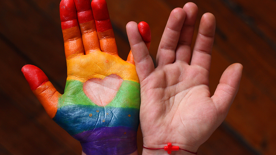 Two hands, one with rainbow painted around a heart shape and the other wearing a faith bracelet