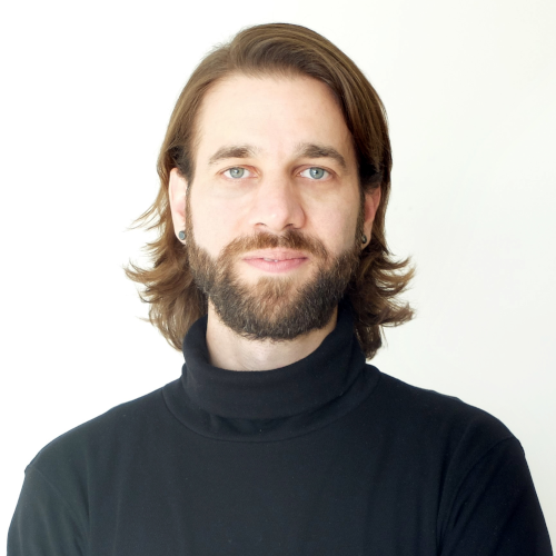 Headshot of a white person with semi-long hair, facial hair, blue eyes and a black turtleneck, standing in front of a white background.