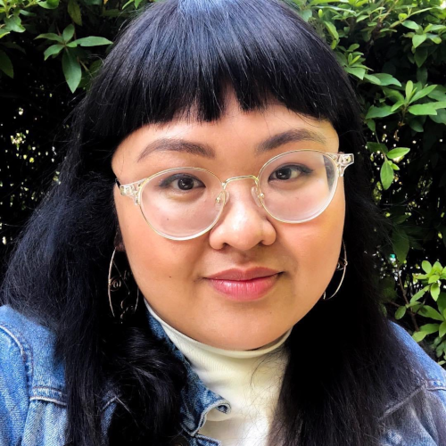 Headshot of an Asian person with black hair and bangs, with dark brown eyes, clear-colored glasses, and a closed-mouth smile. They are wearing a white turtleneck and denim jacket.