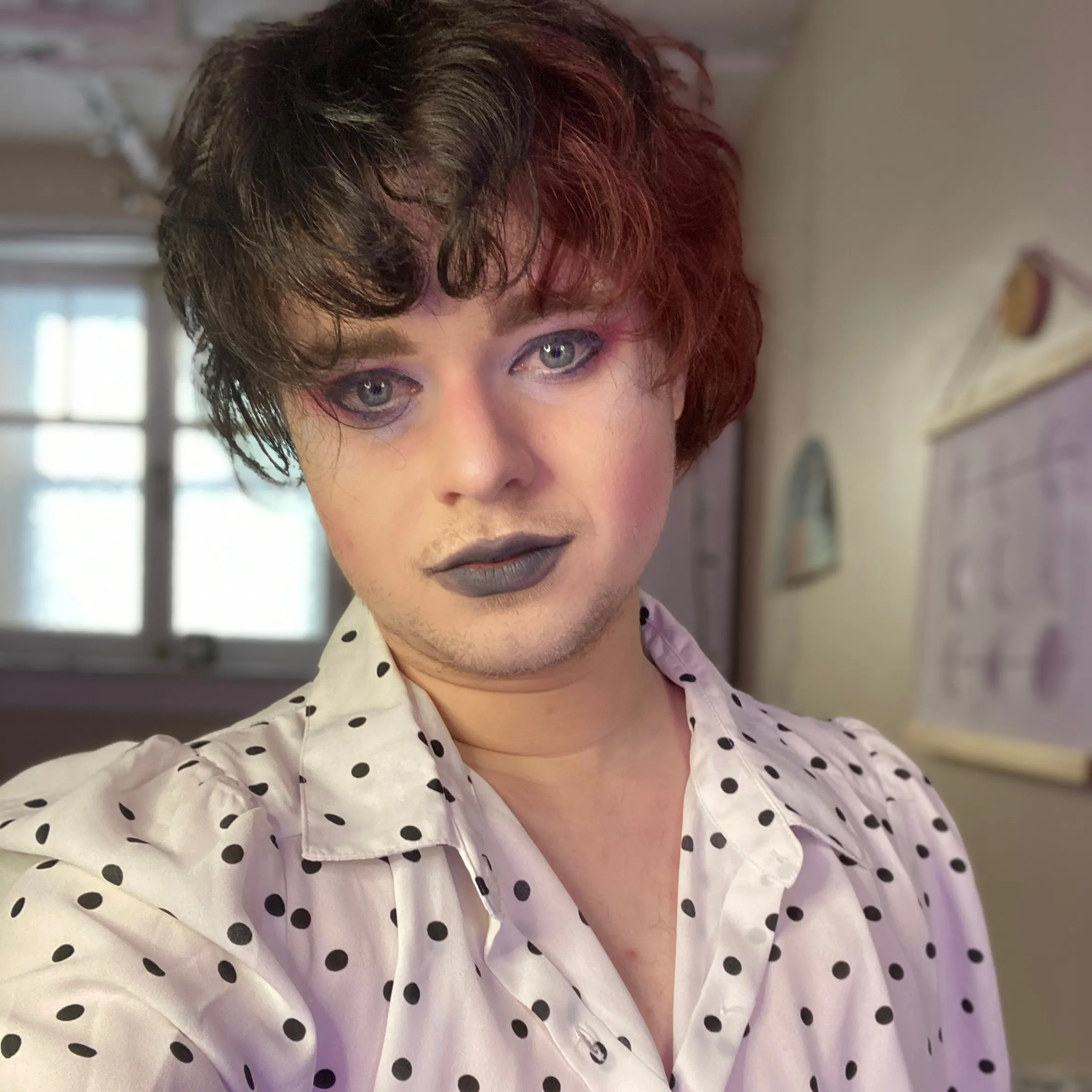 Pictured is a photograph of a white person with medium length, two-toned hair, blue eyes, lightly smiling with cool toned lipstick, wearing a white shirt with black polka dots.]  